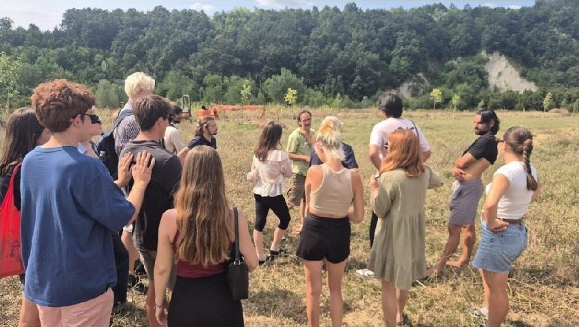 A group gathered in a mown field below wooded hills